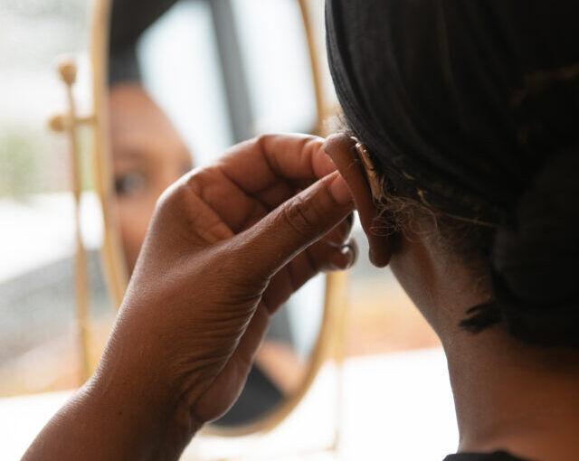 A black woman with short hair sits at a dressing table and puts in a hearing aid
