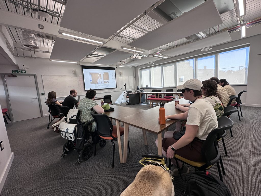 Attendees watching a live stream in a seminar room. There is a wheelchair user and a guide dog in the picture.