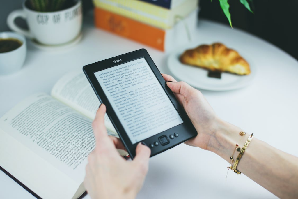 Person holding a Kindle ebook reader with coffee, croissant and print books in background
