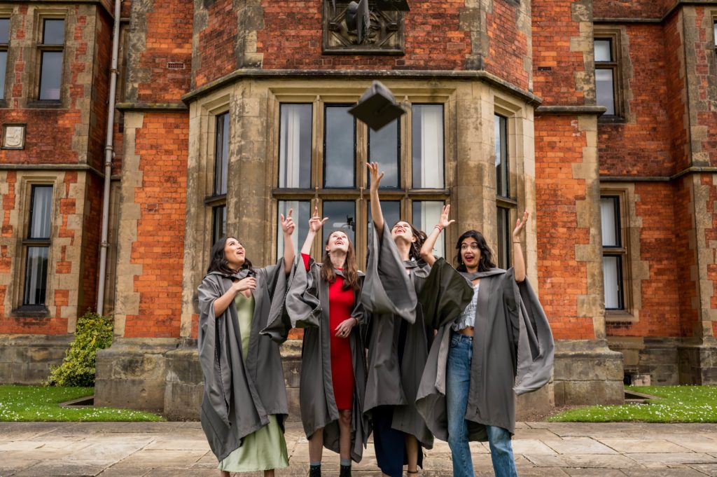 Four graduates in gowns throw their mortarboards into the air outside Heslington Hall.