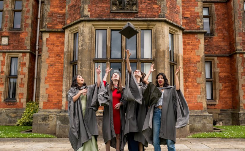Four graduates in gowns throw their mortarboards into the air outside Heslington Hall.