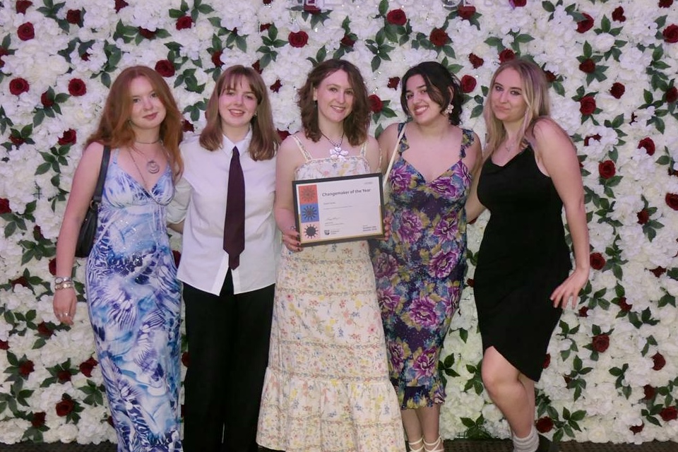 The blog author Susie Symes stands with a group of women from Swap Don't Shop as they pose for a photo with the Changemaker award certificate in front of a floral background